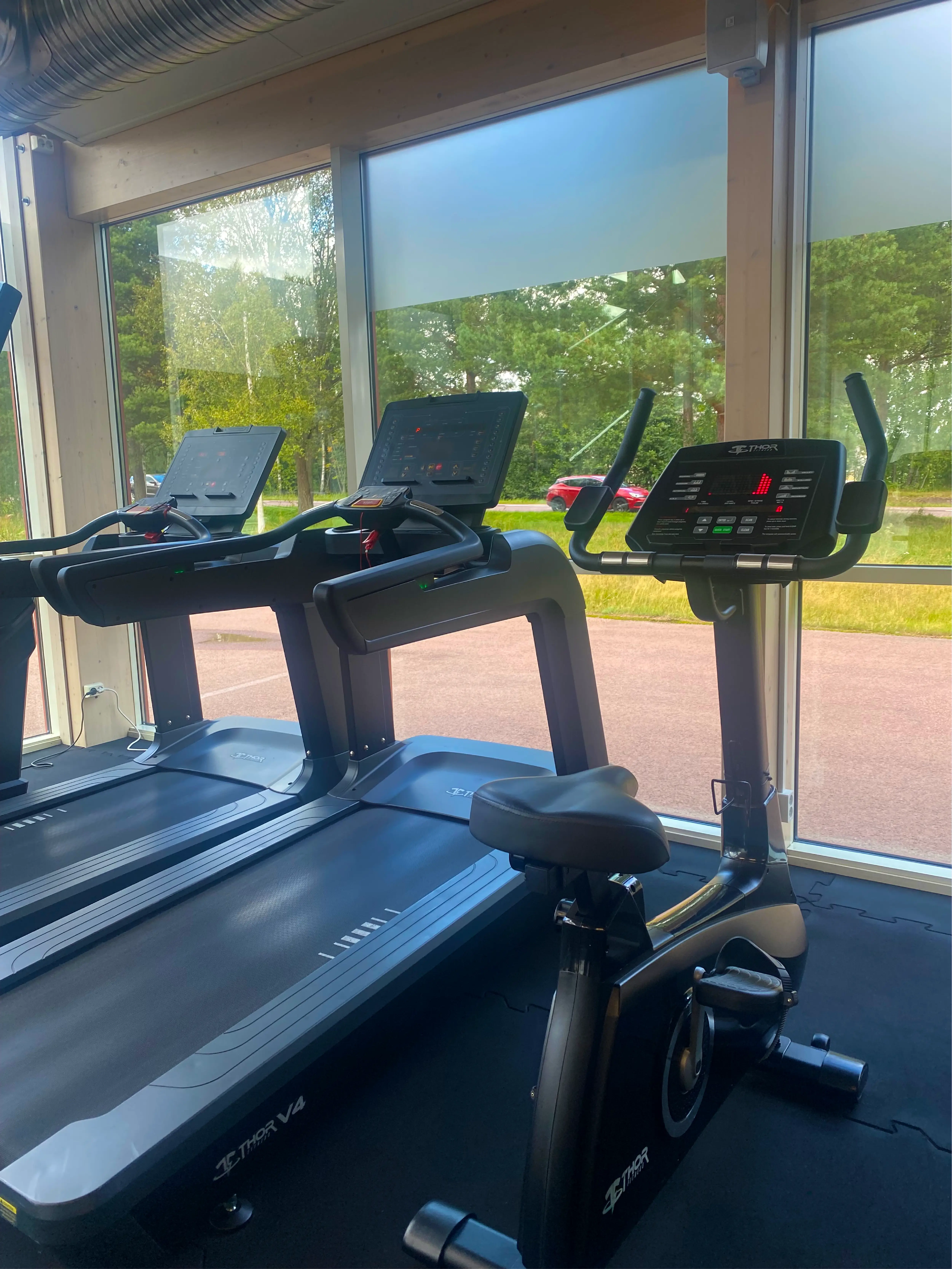 Träningsutrustning vid stora fönster på ett gym – två löpband och en motionscykel med digitala skärmar, med utsikt mot en grönskande omgivning utanför.