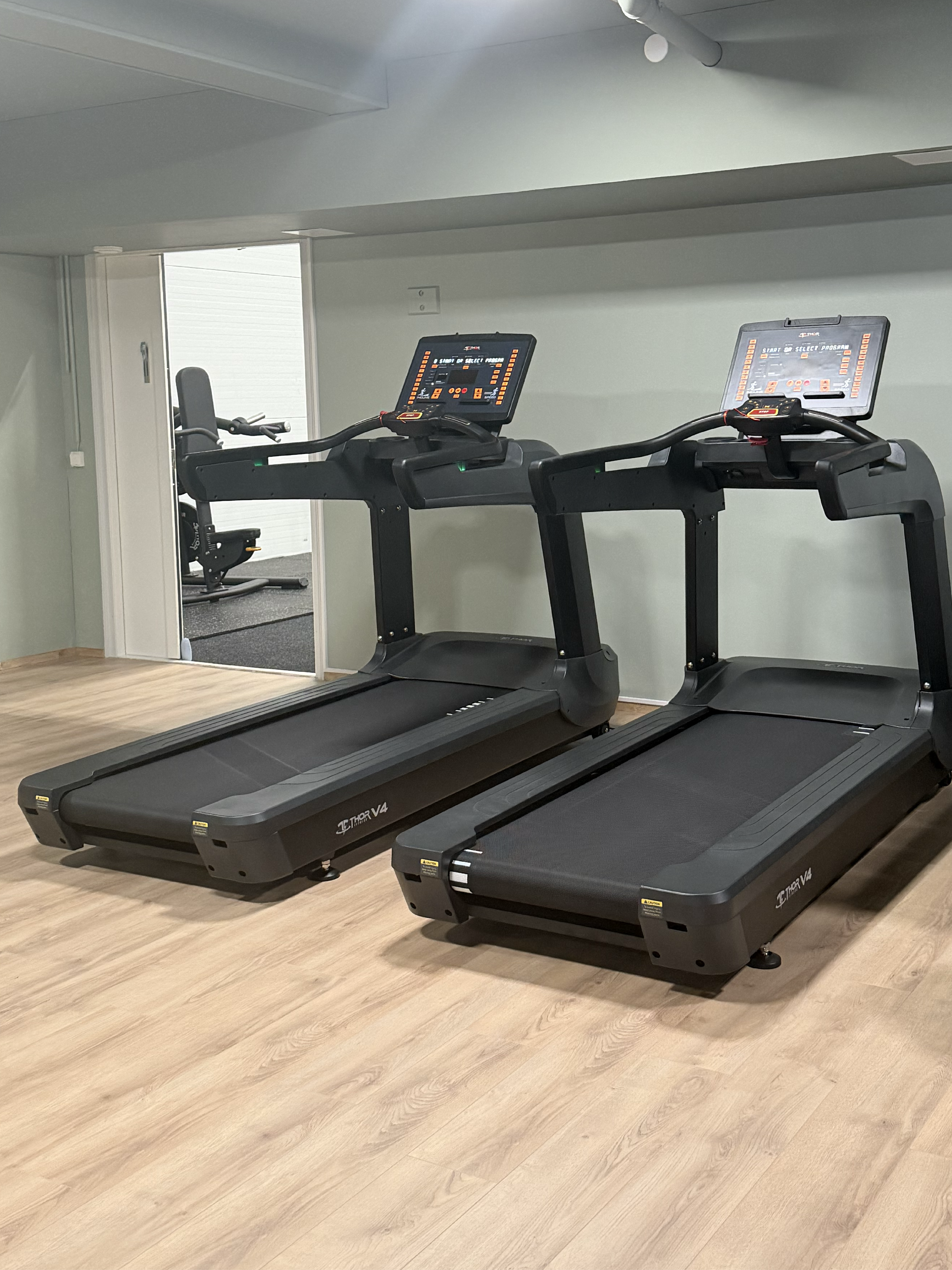 Två löpband står i ett separat rum med trägolv och dämpad belysning, med öppning mot gymmets huvuddel.