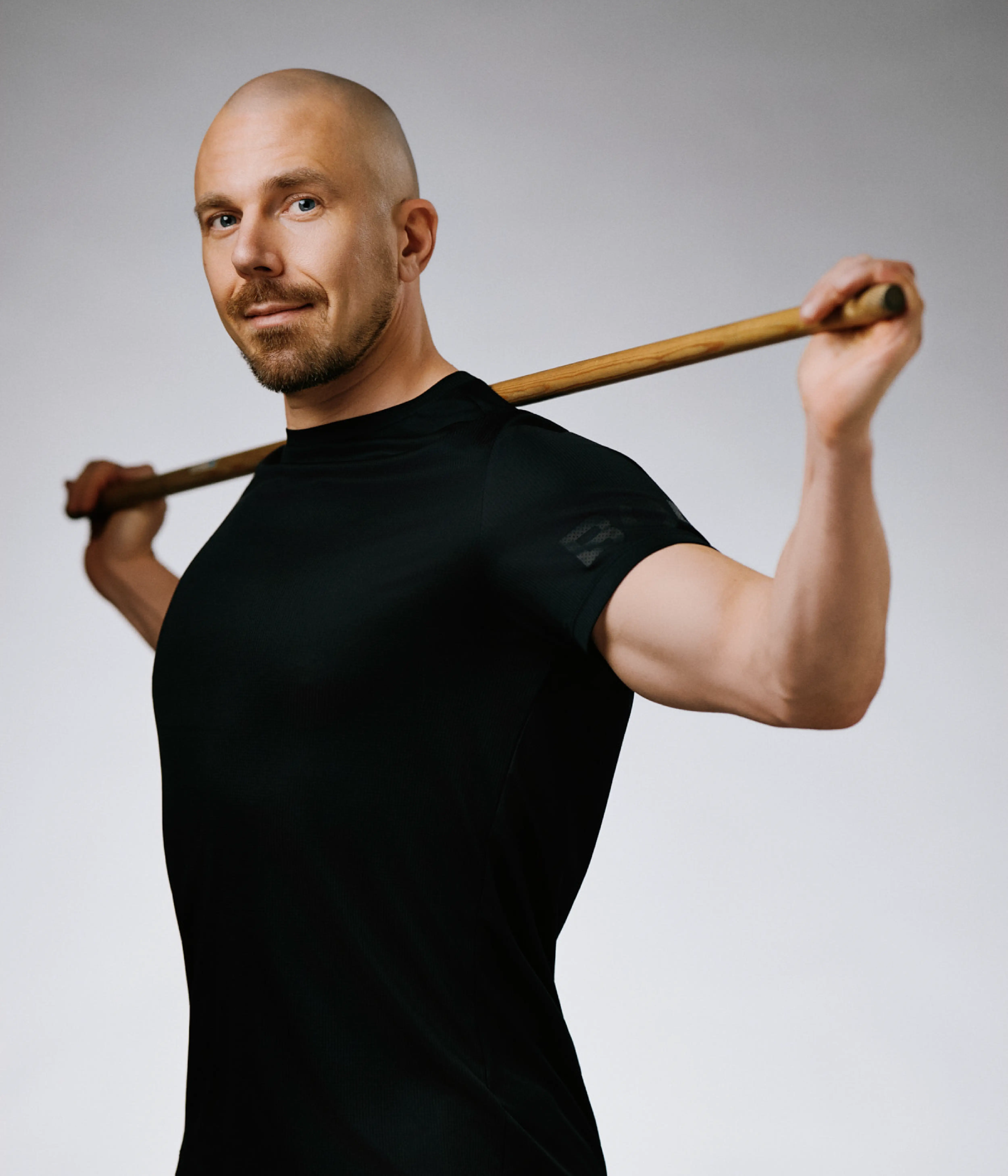 Porträtt av Ken Landström, personlig tränare på Hälsofabriken. Han står i träningskläder med en trästav vilande över axlarna och ser mot kameran med ett lugnt uttryck. Bakgrunden är ljus och neutral.